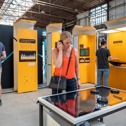 Personen in der Ausstellung Power2Change. Im Hintergrund stehen mehrere Personen an orangefarbenen Exponaten. Im Vordergrund steht eine Person an einem Medientisch und hört etwas über Kopfhörer. 